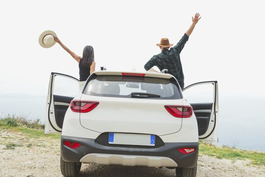 Couple cheers with arms raised while standing by white car overlooking coastal cliff. Bright horizon and open doors evoke sense of freedom and successful road trip journey