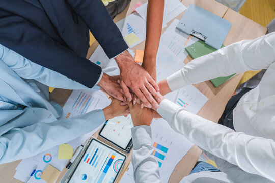 Hands stack together at the center of a busy business desk, symbolizing shared commitment amid ongoing data analysis. This layered overhead composition connects team unity with tangible office product
