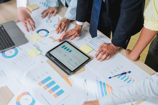 overhead shot of a business team's hands pointing at financial charts on a tablet, surrounded by printed reports and sticky notes. This detailed scene captures collaborative data analysis and strategi