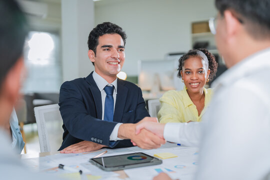 Business partners shake hands across a table covered with charts and documents, sealing a successful agreement. This candid handshake moment captures trust, partnership, and professional deal-making i