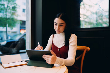 Fototapeta premium profile view of young woman writing with smart stylus on digital tablet screen inside cafe, female student taking online notes from open notebook during distance e-learning course