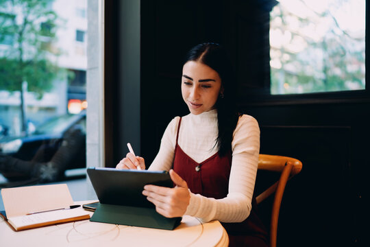 profile view of young woman writing with smart stylus on digital tablet screen inside cafe, female student taking online notes from open notebook during distance e-learning course