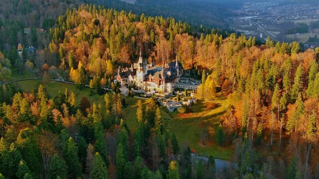 Aerial view of Peles Castle nestled in a dense, autumn-colored forest with golden sunlight illuminating the historic architecture in Romania.