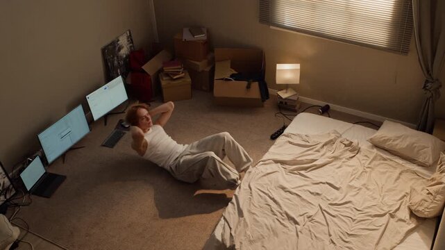 Caucasian young man doing abdominal crunches on floor near computer screens before collapsing tired. Footage ideal for home workout apps, student lifestyle and remote worker wellness projects