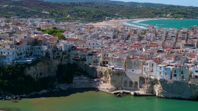 Vieste white old town on limestone cliff aerial 70mm drone tracking shot in Puglia Italy. Lateral flyby along peninsula to San Francesco church over turquoise sea 4K