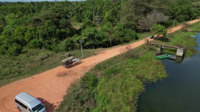 Aerial view of a silver van parked on a dirt road near a lake with lush tropical vegetation and a small boat in Sigiriya, Central Province, Sri Lanka.