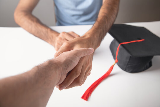 Two people shaking hands with a graduation hat on the table.