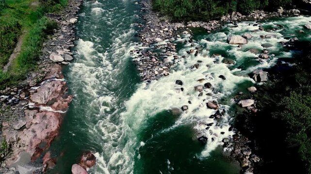High aerial view of a turbulent river dividing around a rocky gravel island. Whitewater currents weave through boulders and exposed stone, illustrating freshwater dynamics, erosion and rugged wilderne