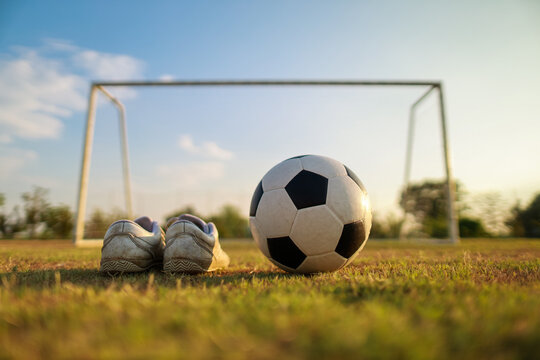 Low angle close up of a classic soccer ball and worn white football boots on a green grass field with a goal net and blue sky background.