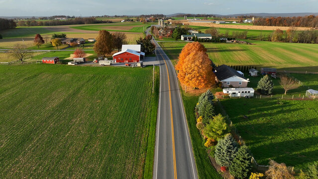 Aerial view of a rural road cutting through farmland with a red barn, autumn foliage, and residential properties in Rapho, Pennsylvania, United States.