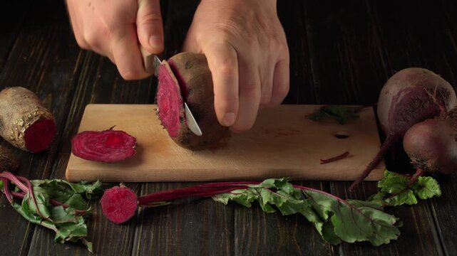 Hands slicing a red beet on a wooden cutting board surrounded by whole beets, beet greens, and freshly cut rounds in a top view kitchen preparation scene