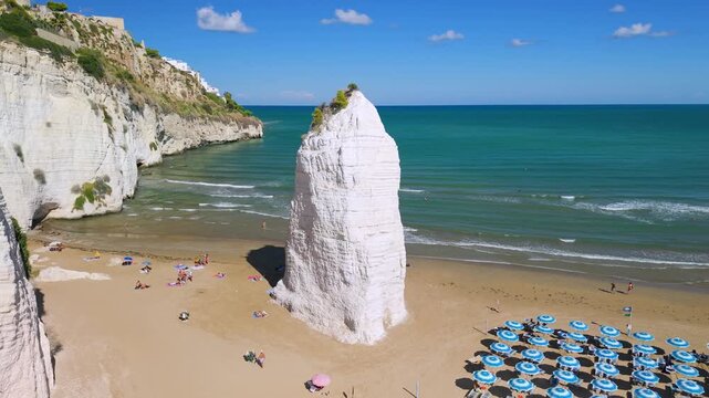 Pizzomunno giant white monolith rock on Scialara beach in Vieste Puglia Italy. Aerial drone rising shot revealing equipped beach and cliff peninsula 4K