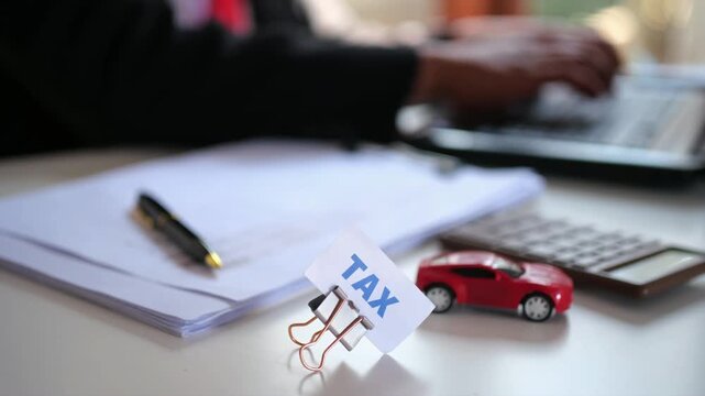 A "TAX" deadline sign and a toy car on a desk, with an accountant typing on a laptop in the background. Financial planning and vehicle tax payment budgeting.
