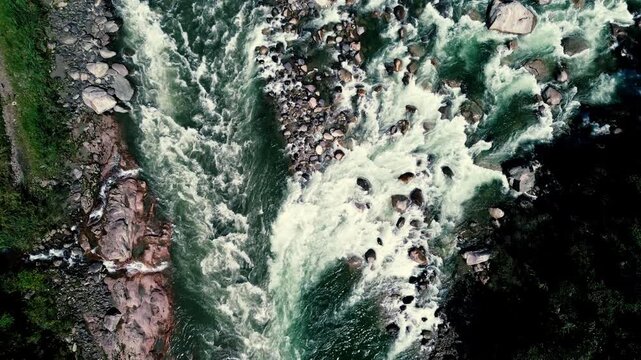 Vertical aerial view of powerful whitewater splitting around a rocky island and scattered boulders. Interlacing currents create a dynamic natural pattern associated with river hydrology, erosion and w