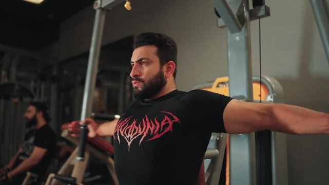 Medium close up low angle shot of a muscular bearded man doing seated machine chest flyes for pectoral muscle isolation and upper body strength training in a gym.