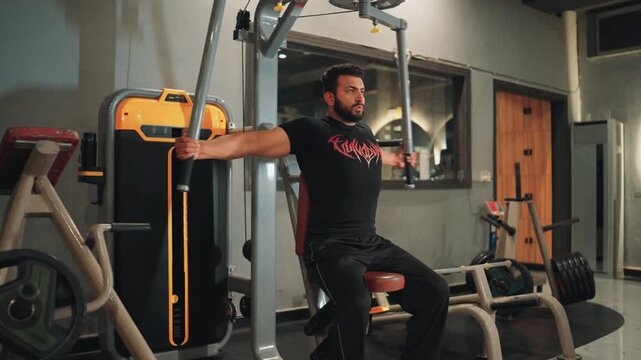 Full body wide view of a muscular bearded man performing seated machine chest flyes for pectoral muscle isolation and upper body strength in a modern gym setting.
