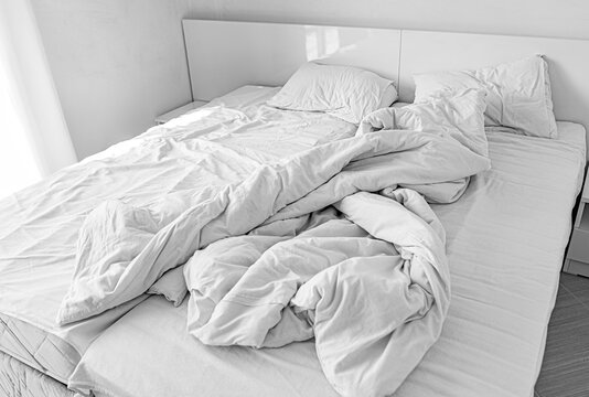 A modern unmade double bed features messy white sheets, a crumpled duvet, and soft pillows in a bright bedroom setting.