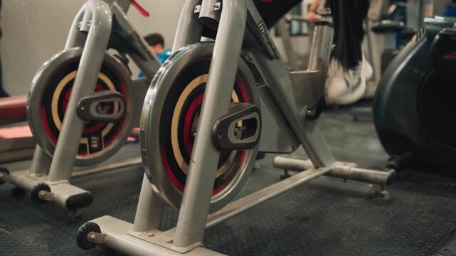 Front view low angle close up shot of a shoe pedaling an indoor exercise spin bike beside a spinning flywheel during an intense gym workout.
