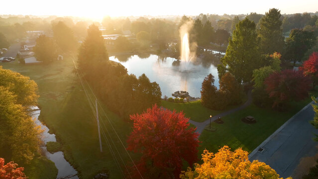 Aerial view of a park with a fountain in a pond surrounded by autumn trees during golden hour in East Lampeter Township, Pennsylvania, United States.