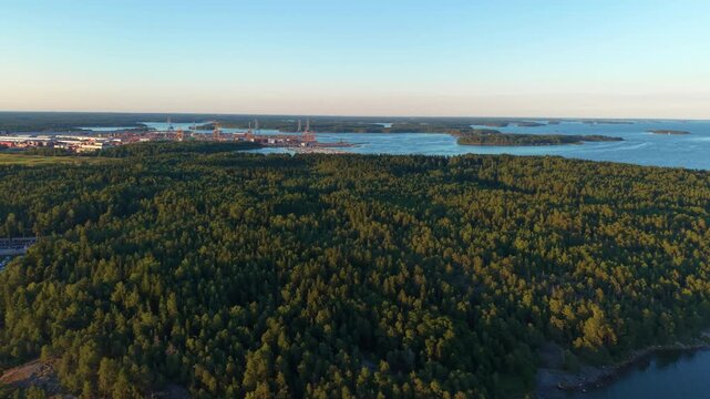 Panoramic view of Finnish Baltic Sea archipelago featuring forested islands, rocky coastline, industrial port with cranes, calm blue water and golden evening light