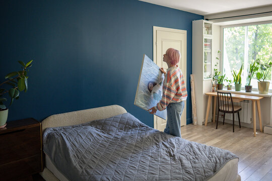 Woman artist holding canvas artwork ready to hang picture over bed on wall in bedroom, creative female painter stands beside bed preparing to place painting in room, wall decor and cozy living space.