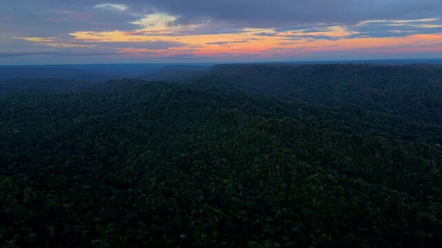 Aerial view across expansive tropical rainforest hills beneath an orange and pink sunset. The remote wilderness landscape evokes tranquillity, biodiversity, natural beauty and forest conservation.