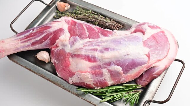 Raw leg of lamb with garlic and fresh rosemary on a rectangular metal tray rotating against a plain white studio background.