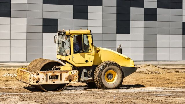 A yellow roller compacts dirt on a construction site next to a building with a gray and black facade. Workers are present in the background