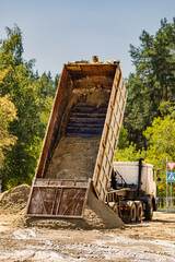 A dump truck tilts its bed to release a load of sand at a construction site. The area features trees and a road sign, with clear visibility © Anoo
