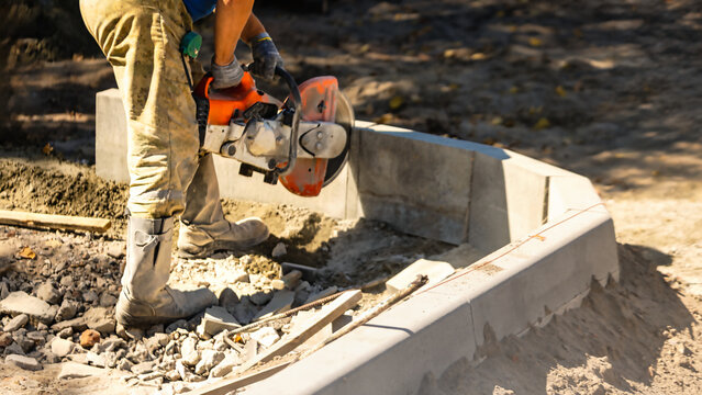 A worker uses a concrete saw to cut a slab on a construction site. The ground is covered with rocks and dust. Tools are nearby, and the work is ongoing