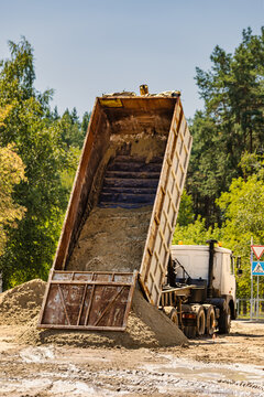A dump truck tilts its bed to release a load of sand at a construction site. The area features trees and a road sign, with clear visibility