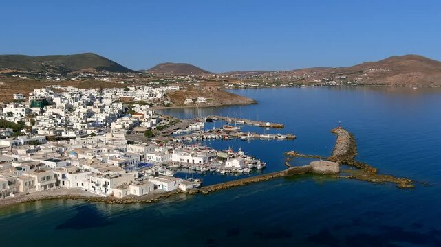 Aerial view of the white buildings of a coastal town with a harbor filled with boats and clear blue sea in Islands, Greece.