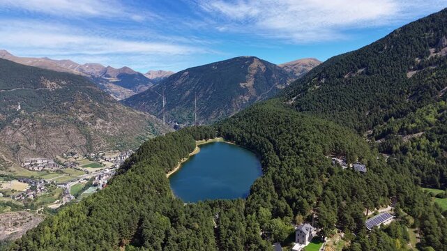 Aerial view of Lake Engolasters nestled among dense pine forests in Andorra, with Pyrenees mountain peaks, valley settlements and blue sky in background