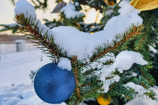 A Christmas tree stands covered in snow with colorful ornaments. The scene shows winter joy as people celebrate the New Year in a festive environment