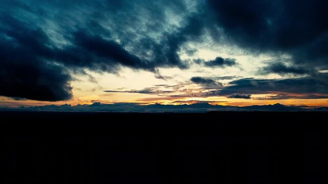 Dark storm clouds stretch above a glowing orange sunset and silhouetted mountain horizon. The atmospheric sky creates a dramatic natural background associated with changing weather, twilight and power