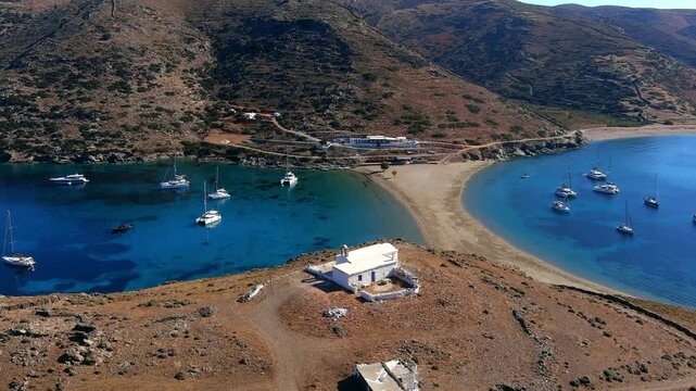 Aerial view of the double-sided Kolona Beach with a white church on the isthmus, surrounded by turquoise waters and anchored sailboats in Islands, Greece.