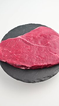 Single raw lean beef steak on a round black slate serving board rotating against a clean white background in vertical close-up.