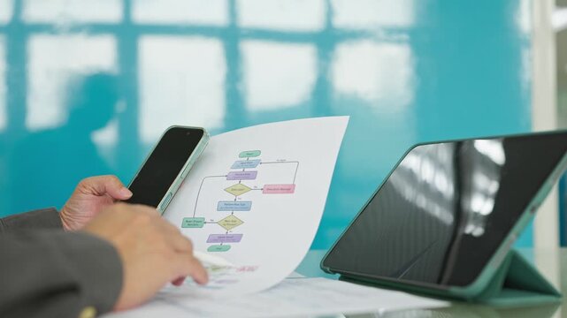 Professional holding smartphone reviews printed flowchart diagram document beside digital tablet computer at office desk, planning software algorithm workflow development process.