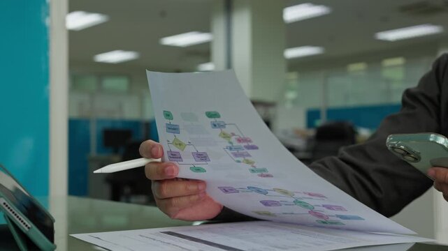 Professional programmer holding printed algorithm flowchart document holding stylus pen reviewing software logic inside corporate office workplace environment.