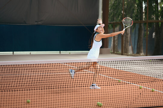 Young Asian Woman Playing Tennis on Clay Court, Swinging Racket in Dynamic Action Shot