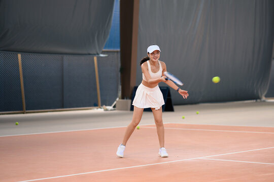 Young Asian Woman Playing Tennis on Indoor Court, Hitting Forehand Shot in White Sportswear