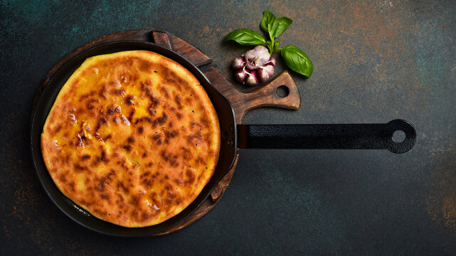 Traditional roasted round pie baked inside black frying pan top view. Tasty golden cheese flatbread surrounded by garlic and chili peppers. Gourmet homemade meal recipe.