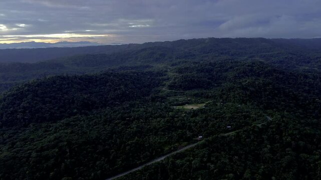 Wide aerial view of a winding road crossing densely forested tropical hills beneath dark storm clouds. A small clearing and scattered buildings suggest remote rural infrastructure surrounded by wilder