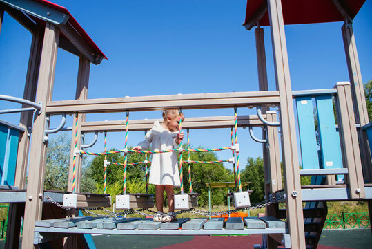 A girl in a white dress plays on the playground, climbs a rope ladder, and on the bars at the top of the slide.