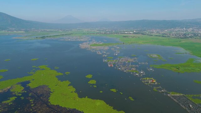 Aerial view of Lake Rawa Pening with fish farming structures and lush greenery surrounded by mountains in Waduk Rawapening, Central Java, Indonesia.
