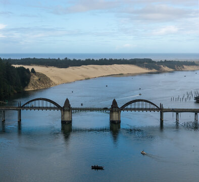 Aerial view of the Siuslaw River Bridge spanning the river with sand dunes in the background and small fishing boats on the water in Florence, Oregon, United States.