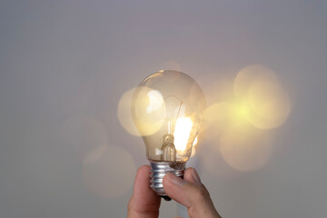 Person's hand grips lit incandescent bulb, glowing light bulb against soft bokeh background. © jittawit21