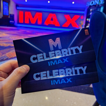 Hand holding M Celebrity IMAX movie tickets in front of large red glowing neon IMAX logo background in modern cinema theater hall on September 28, 2026 in Bangkok, Thailand.