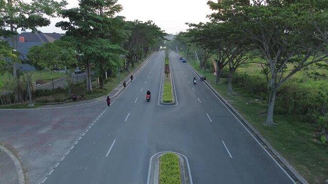Aerial view of a wide asphalt road with a lush green median and tall trees in a peaceful suburban neighborhood, featuring people jogging and