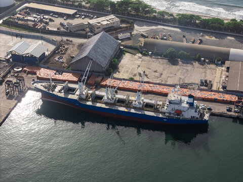Aerial view of a cargo ship docked at the industrial port with stacks of orange cargo containers and warehouses nearby in Bajos de Haina, San Cristobal Provinc, Dominican Republic.